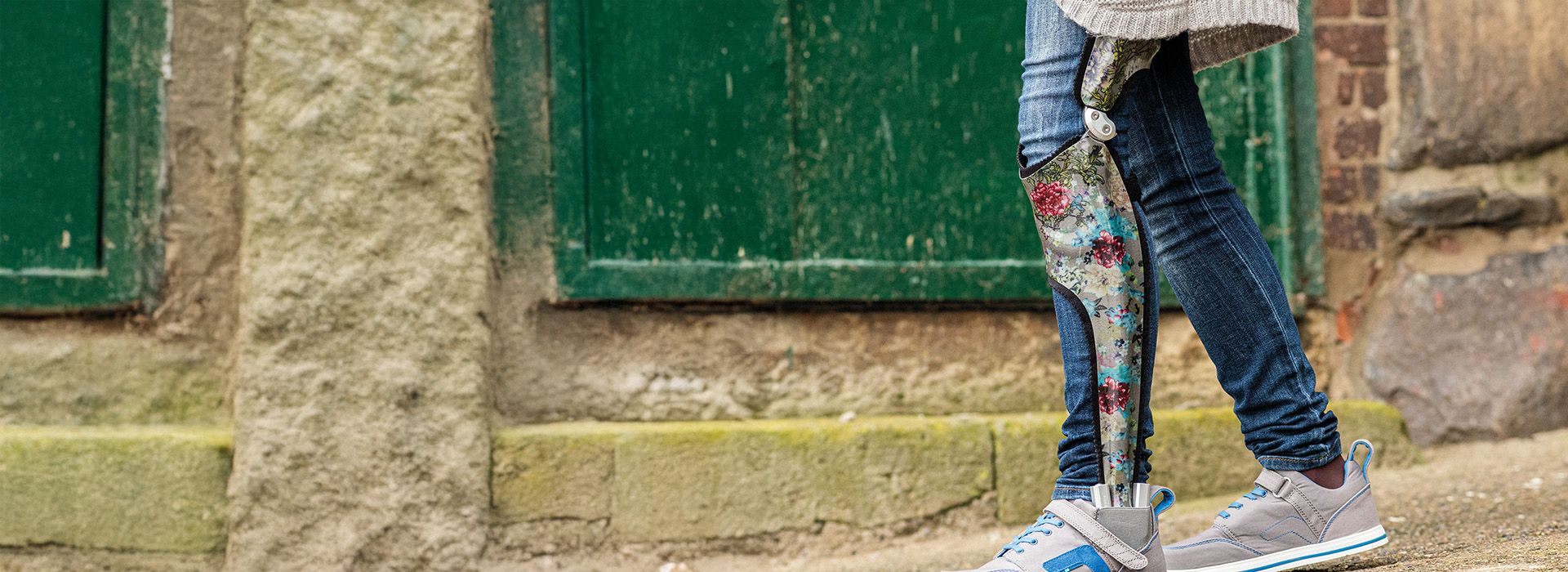 Bein mit bunter Prothese, Jeans und grauen Sportschuhen vor einer alten Steinmauer mit grünen Fensterläden